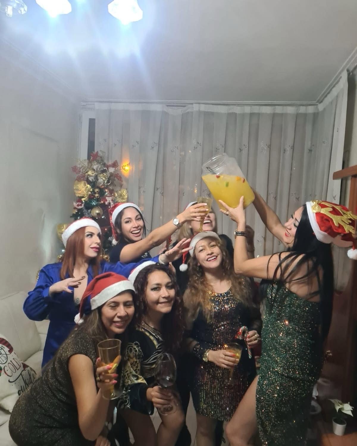 Grupo de mujeres con gorros navideños celebrando con bebidas frente a un árbol de Navidad